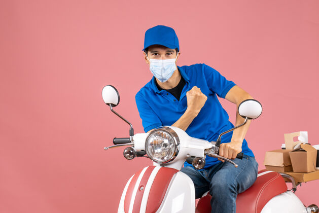摩托车俯视图自豪的送货员戴着医用面具 戴着帽子坐在粉彩桃色背景的踏板车上坐摩托车背景