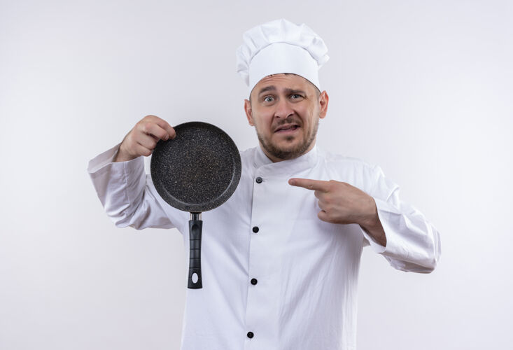 帅哥印象深刻的年轻帅哥厨师长制服举行 并指着煎锅隔离在白色的墙壁拿着指着薯条