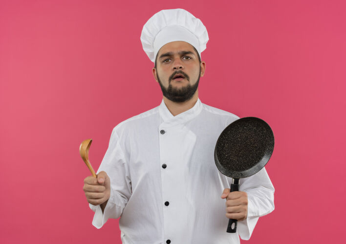 勺子困惑的年轻男厨师穿着厨师制服 手里拿着煎锅和汤匙 被隔离在粉红色的墙上年轻平底锅困惑