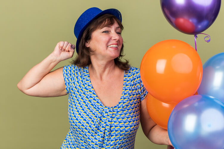 气球快乐兴奋的中年妇女戴着派对帽 手里拿着一堆五颜六色的气球 开心地笑着站在绿色的墙上庆祝生日派对乐趣站立微笑