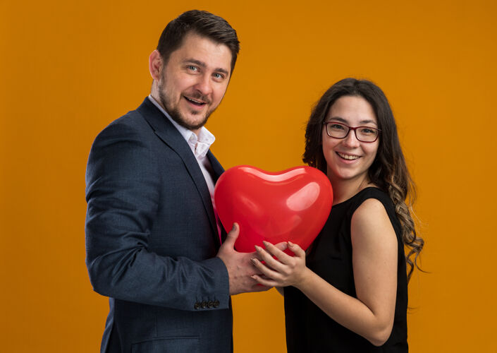 男人幸福而美丽的情侣们手拿心形的红色气球 在橙色的墙上拥抱庆祝情人节女人礼物拥抱