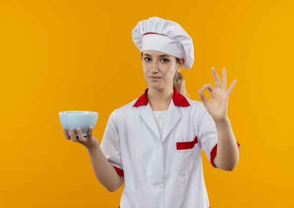 碗自信的年轻漂亮厨师 身着厨师制服 拿着碗 在橙色的墙上贴着“一切正常”的牌子拿着厨师好