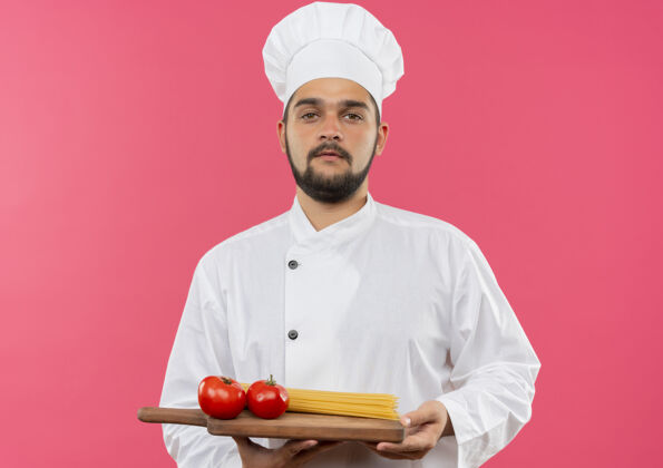 男性自信的年轻男性厨师身着厨师制服 拿着砧板 上面有西红柿和意大利面 隔离在粉红色的墙上制服意大利面厨师