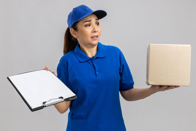 制服身穿蓝色制服 头戴鸭舌帽的年轻女送货员站在白色的墙上 手里拿着纸板箱和剪贴板 困惑地看着一旁站在一边举行年轻