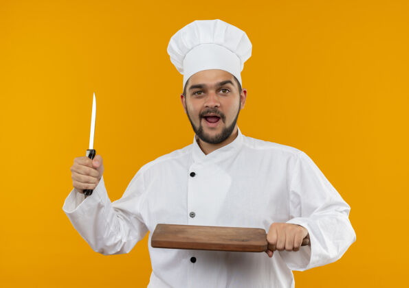 烹饪印象深刻的年轻男厨师在厨师制服持刀和砧板隔离在橙色的墙上板年轻持有