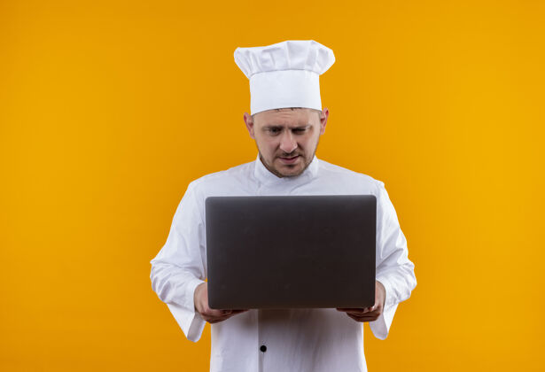 持有迷茫的年轻帅哥厨师穿着厨师制服拿着笔记本电脑看着隔离在橙色墙上帅气笔记本电脑制服