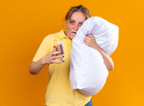 抱着不健康的女人穿着黄色衬衫 患流感和感冒 感觉不舒服 抱着枕头 拿着药片和一杯水站在橙色的墙上拥抱站着女人