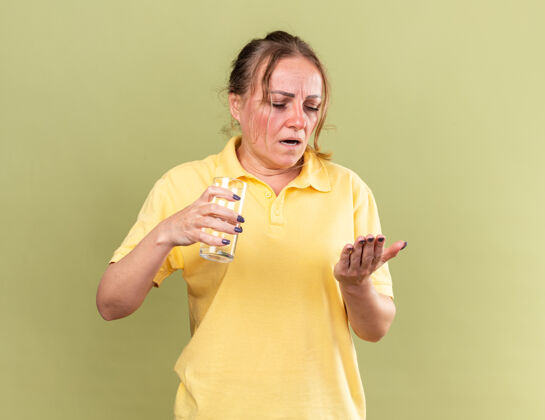 可怕不健康的黄衬衫女人站在绿色的墙上 手里拿着一杯水和药片 正准备吃药 患流感和感冒 感觉很糟糕站着不健康女人