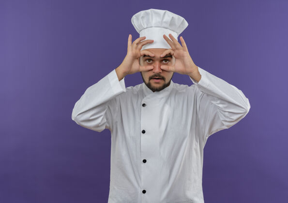 制服令人印象深刻的年轻男厨师在厨师制服做看手势用手作为望远镜隔离在紫色的墙上烹饪男性望远镜