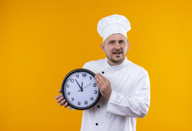 厨师快乐的年轻帅哥厨师穿着厨师制服 拿着橙色墙上孤立的时钟帅气年轻制服