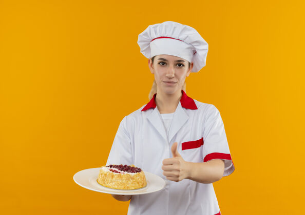 厨师自信的年轻漂亮厨师 身着厨师制服 拿着一盘蛋糕 在橙色的墙上孤立地竖起大拇指制服拿着年轻