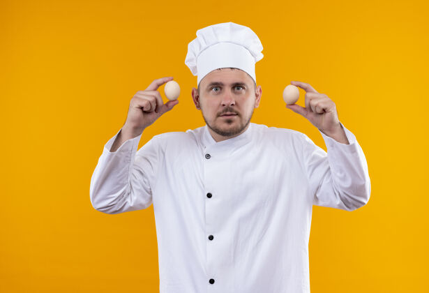 印象深刻年轻帅气的厨师穿着厨师制服 手里拿着隔离在橙色墙上的鸡蛋手握厨艺制服