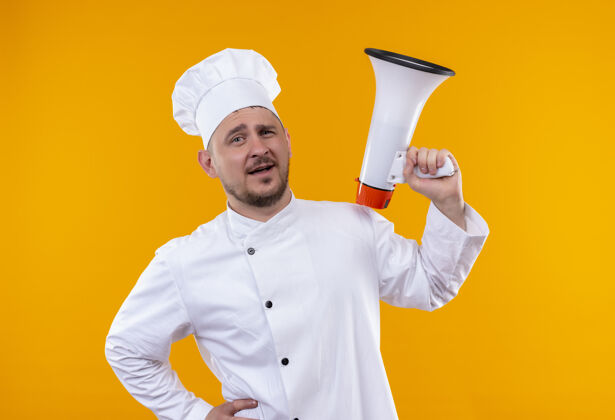 自信自信的年轻帅哥厨师穿着厨师制服 在孤立的橙色墙上拿着扬声器持枪年轻厨师