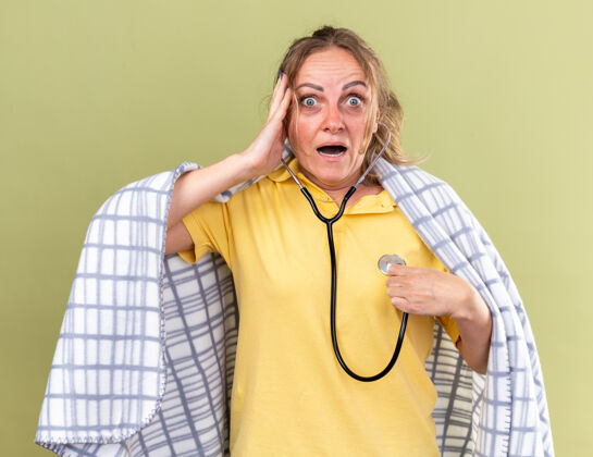 担心不健康的女人裹着毯子 感觉不舒服 患流感和感冒 站在绿色的墙上 用听诊器听心跳 看起来很担心感觉不健康心跳