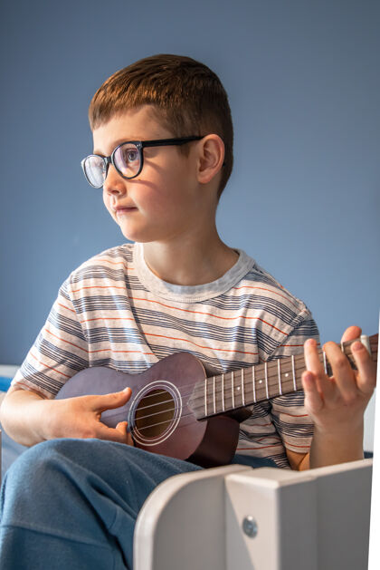 年轻一个戴眼镜的可爱男孩在家里 在他的房间里学着弹四弦琴床孩子经典