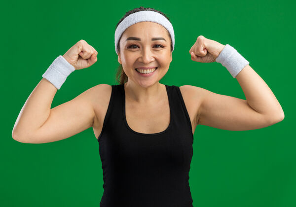 健康戴着头带和臂章的快乐的年轻健身女士微笑着自信地举起拳头站在绿色的墙上头带微笑臂章