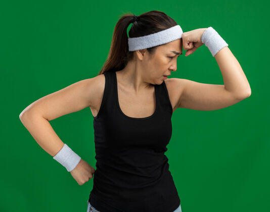 头带戴着头巾和臂章的年轻健身女士站在绿色的墙上 一边沉思一边思考健康臂章旁白