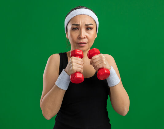 女人戴着头带和哑铃做运动的年轻健身女士紧张而自信地站在绿色的墙上头带健身哑铃