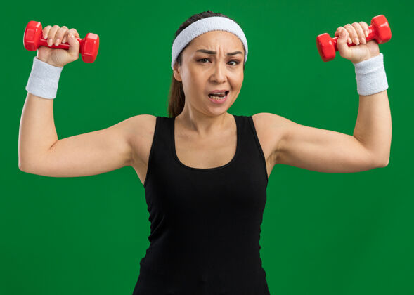 年轻戴着头带和哑铃做运动的年轻健身女士紧张而自信地站在绿色的墙上健身锻炼自信
