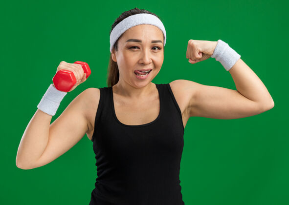 锻炼戴着哑铃的头带做练习的年轻健身女士举起拳头 露出二头肌微笑自信地站在绿色的墙上自信二头肌微笑