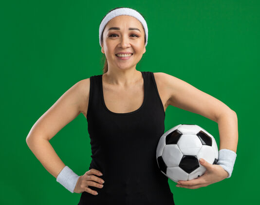 袖带戴着头带和臂章的快乐的年轻健身女士 面带微笑地举着足球站在绿色的墙上健身女人抱