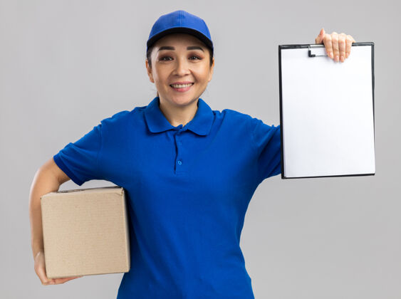 女人身穿蓝色制服 戴着帽子的年轻送货妇女站在白色的墙上 手里拿着一个纸板箱 上面有空白页的剪贴板 微笑着自信地站着交货页面制服