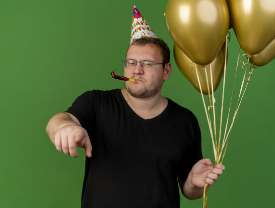 吹自信的成年斯拉夫人戴着眼镜 戴着生日帽 手里拿着氦气球吹着派对哨子 指着摄像机眼镜聚会帽子