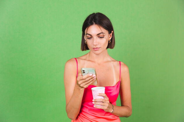 信息时尚女人穿着粉色丝质连衣裙孤立在绿色上开心地笑着看手机屏幕上的消息新闻情感快乐在线