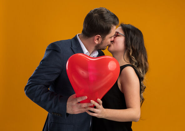 亲吻幸福而美丽的情侣 心形红色气球的男女 在橙色的墙上拥抱亲吻庆祝情人节女人情侣气球