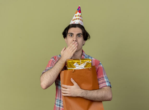 穿戴着生日帽的白种人帅哥震惊地把手放在嘴边 手里拿着纸购物袋里的礼盒帽子生日盒子