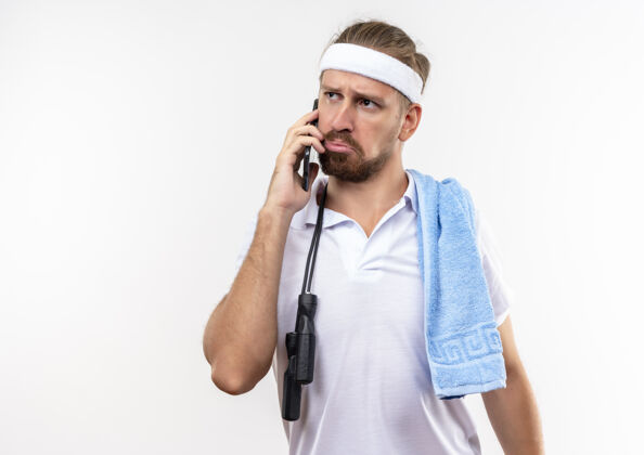 运动困惑的年轻英俊的运动型男子戴着头带和腕带 用毛巾和跳绳在肩上打电话 看着隔离在白色墙壁上的一面 还有复制空间壁板肩绳子