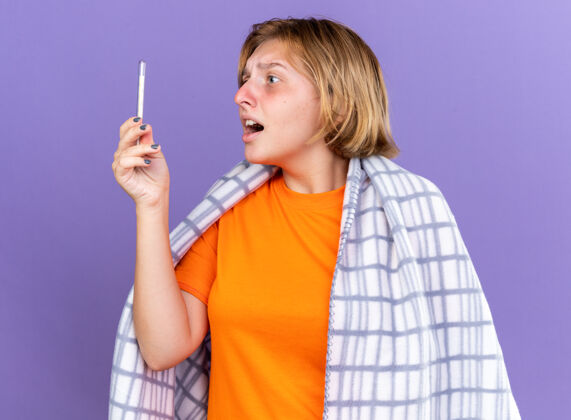 使用不健康的年轻女子裹着温暖的毯子 感觉不舒服 患流感 发烧 用温度计测量体温 站在紫色的墙上 看起来很担心暖和年轻生病