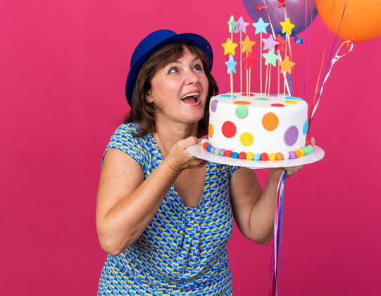 蛋糕快乐快乐的中年妇女戴着派对帽 手里拿着五颜六色的气球 手里拿着生日蛋糕 面带微笑 站在粉色的墙上庆祝生日派对微笑女人生日