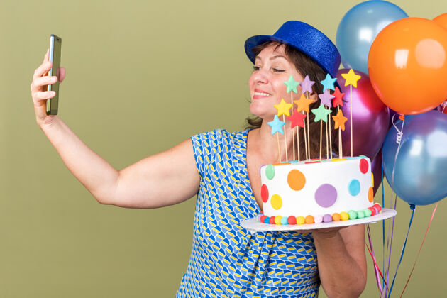 自拍快乐的中年妇女戴着派对帽 手里拿着一堆五颜六色的气球 手里拿着生日蛋糕 用智能手机自拍 站在绿色的墙上庆祝生日派对气球女人年龄