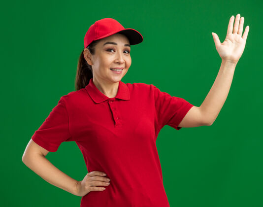 微笑身穿红色制服 头戴帽子的年轻送货员快乐而积极地举手微笑着站在绿色的墙上站立年轻积极