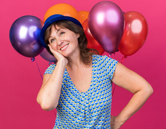 中间快乐快乐的中年妇女 戴着派对帽 手持五颜六色的气球 面带微笑 站在粉色的墙上庆祝生日派对立场脸女人