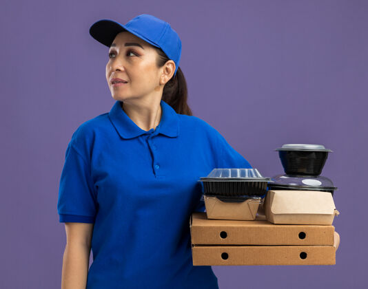 脸身穿蓝色制服 头戴鸭舌帽的年轻送货员站在紫色的墙上 手里拿着披萨盒和食品包 面带微笑地看着一旁微笑女人送货