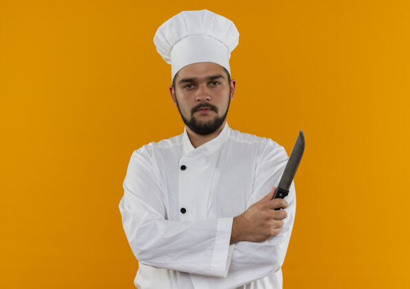 年轻自信的年轻男厨师身着厨师制服 站着封闭的姿势 拿着隔离在橙色墙上的刀制服烹饪站立