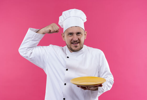 制服快乐的年轻帅哥厨师穿着厨师制服拿着盘子在粉红色的墙上做着强烈的孤立的手势强壮盘子抱着
