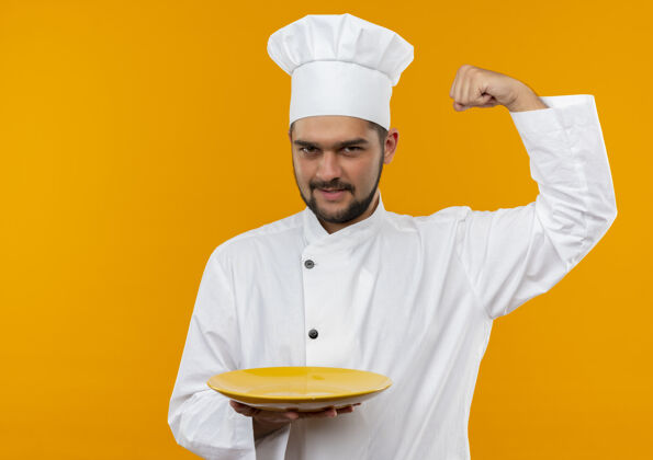 自信自信的年轻男厨师身着厨师制服 拿着空盘子 在橙色的墙上做着强烈的孤立的手势盘子姿态抱着