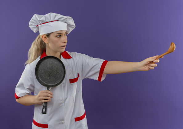 年轻年轻漂亮的厨师穿着厨师制服 拿着煎锅望着一边 伸出勺子 孤立地站在紫色的墙上制服印象勺子