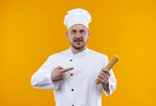 意大利面穿着厨师制服的年轻帅哥厨师手拿意大利面 指着隔离在橙色墙上的意大利面指着烹饪印象深刻