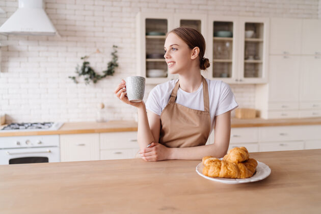 快乐穿着围裙 咖啡和羊角面包的年轻漂亮女人在厨房里享受她的早晨手咖啡渣欢乐
