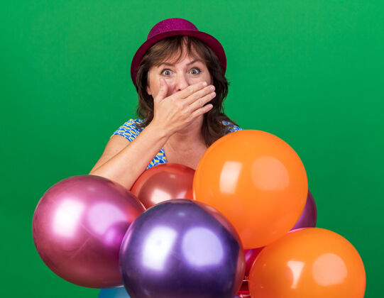 帽子中年妇女戴着派对帽 手里拿着五颜六色的气球 惊讶地捂着嘴 站在绿色的墙上庆祝生日派对气球女人手