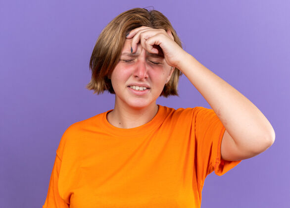 女人不健康的年轻女子穿着橘色t恤 站在紫色的墙上摸头感到头晕感冒 感觉很糟糕站而可怕