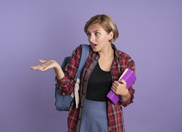 女孩震惊的年轻斯拉夫学生女孩穿着背包拿着书和笔记本看着并指着旁边紫色指向学生