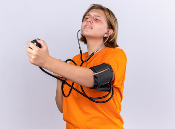 年轻不健康的年轻女子穿着橙色t恤 感觉不舒服 用血压计测量血压 站在白色的墙上 看起来很担心感觉血压计压力