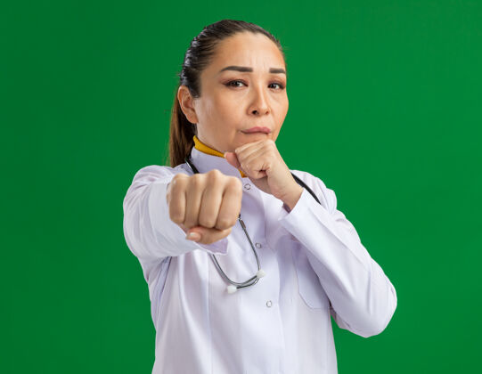 站一位年轻的女医生 身穿白药衣 脖子上戴着听诊器 严肃的脸像一个拳击手 紧握拳头站在绿色的墙上姿势外套年轻