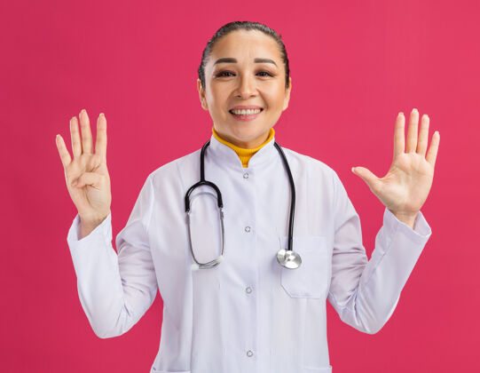 站身穿白色药衣 脖子上戴着听诊器的年轻女医生微笑着 手指站在粉红色的墙上 显示着九号医生围着表演
