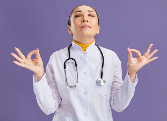 放松年轻的女医生穿着白色药衣 脖子上戴着听诊器 闭着眼睛做着冥想的手势 站在紫色的墙上放松手指做女人